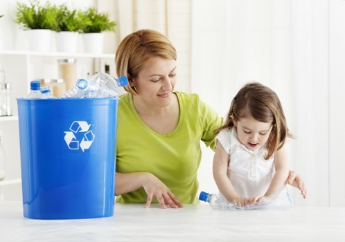 A woman with short, light brown hair wearing a neon green top is seated at a table indoors, assisting a young girl with shoulder-length brown hair dressed in a white top. They are both focused on a clear plastic bottle that the girl is holding and examining carefully. To the left of the woman, there is a large blue recycling bin made of plastic with a white recycling symbol printed on it, filled with various crumpled plastic bottles and containers, some of which are partially visible and spilling slightly over the edge. The background includes a white shelving unit with multiple small potted plants and white decorative objects, set against a bright, well-lit room with sheer white curtains allowing diffuse natural light to fill the space. The scene emphasizes waste sorting and recycling activities, reflecting environmentally conscious practices typical of local rubbish removal services like Garden Clearance Barbican, situated within the London area, particularly around the Barbican district.