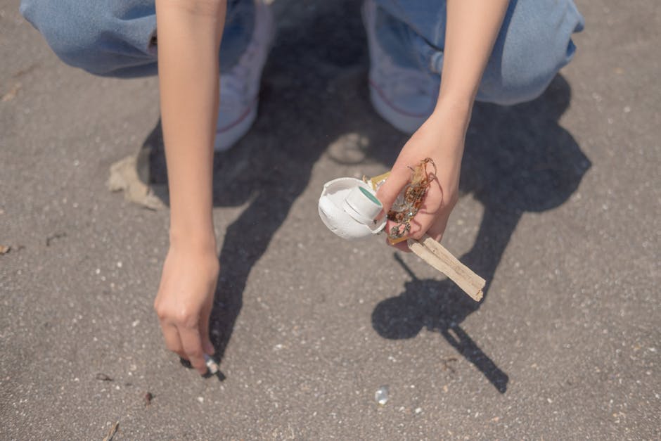 A person wearing a light blue t-shirt, dark shorts, and white sneakers is seen using a litter picker tool to collect small debris from a rocky shoreline. They are holding a white trash bag with a pink