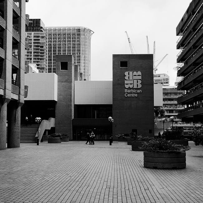 Close-up view of the corner of a modern building with a textured, dark brown, concrete or stone overhang supported by a large, rounded pillar. Part of a sign reading 'Shakespeare' is visible beneath t