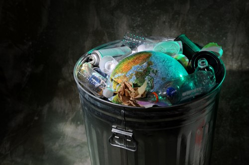 A metal rubbish bin with a hinged lid, filled with various discarded items including crumpled brown paper, plastic bottles, and containers, with a small illuminated globe placed among the waste. The background is dark and textured, emphasizing the contents of the bin and the reflective surface of the metal. This image illustrates general waste suitable for rubbish removal services, highlighting typical household or office rubbish such as plastics and paper, in a setting that could be part of a commercial or domestic environment, possibly in an urban area. The presence of a globe among the waste underscores the importance of waste separation and environmental considerations relevant to services offered by Garden Clearance Barbican in London postcode areas, aligning with rubbish removal and recycling efforts in central London locations.