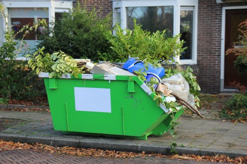 A large bright green skip bin filled with a variety of garden and household waste materials, positioned on a paved driveway in front of a residential property. The contents include branches, leafy plant debris, cardboard boxes, and plastic items, all loosely piled with some extending over the edges of the bin. The skip is situated directly on the pavement, with a curb visible on the street side. In the background, there is a brick house with white-framed windows, some greenery and trees surrounding the property, indicating a suburban neighbourhood. The scene appears to be in daylight, with natural light illuminating the waste materials and the surrounding environment. The presence of the skip suggests a professional rubbish removal service, such as Garden Clearance Barbican, handling garden waste collection in the area, which may be within the postcode or nearby town context.