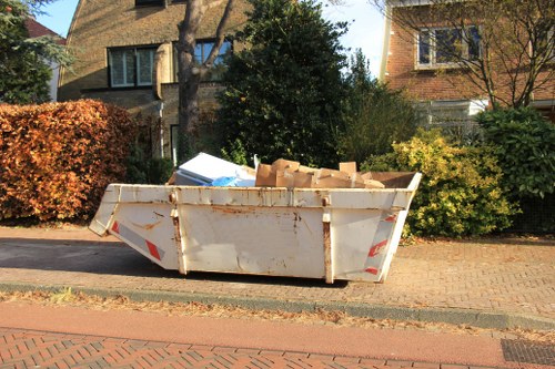 A large, rectangular steel skip with a weathered, off-white finish and red reflective strips, positioned on a paved driveway in front of a residential building. The skip is partially filled with various discarded cardboard boxes of different sizes and light brown color, some protruding over the edge. Surrounding the skip are neatly maintained bushes and trees, with a mix of green and autumnal foliage, and neighboring houses visible in the background. The scene suggests a waste collection or rubbish removal process, typical of garden clearance services in the Barbican area. The overall environment appears calm, with natural daylight illuminating the scene, emphasizing the textures of the cardboard and the surface of the skip, aligned with professional rubbish removal practices by Garden Clearance Barbican.